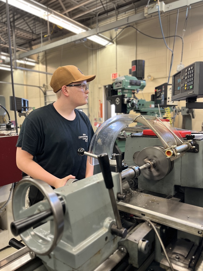 A person wearing a black t-shirt, safety glasses, and a brown cap operates a lathe machine in a workshop. The lathe has a transparent protective cover and visible control knobs. The background includes additional industrial equipment, emphasizing a safe and professional technical environment.