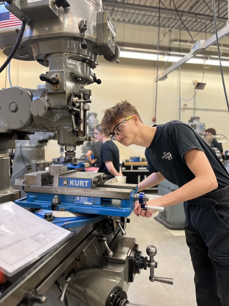 A person wearing safety glasses operates a milling machine equipped with a "KURT" vise in a workshop or manufacturing lab. Other individuals are visible working in the background, suggesting an educational or professional setting focused on machining or manufacturing skills.