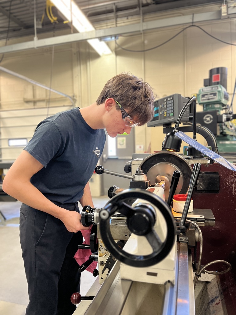 A person wearing safety glasses operates a lathe machine in a workshop, machining a cylindrical object. The workspace contains various tools and equipment, emphasizing hands-on technical work and adherence to industrial safety practices.