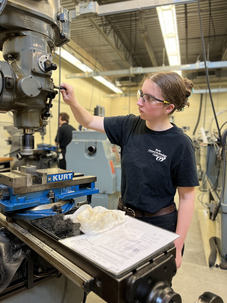 A person wearing safety glasses and a black t-shirt with the words "Kurt" and "Engineering" operates a milling machine in a workshop. The machine features a vise labeled "KURT" and various control components. Additional industrial equipment is visible in the background, highlighting a hands-on engineering or manufacturing environment.