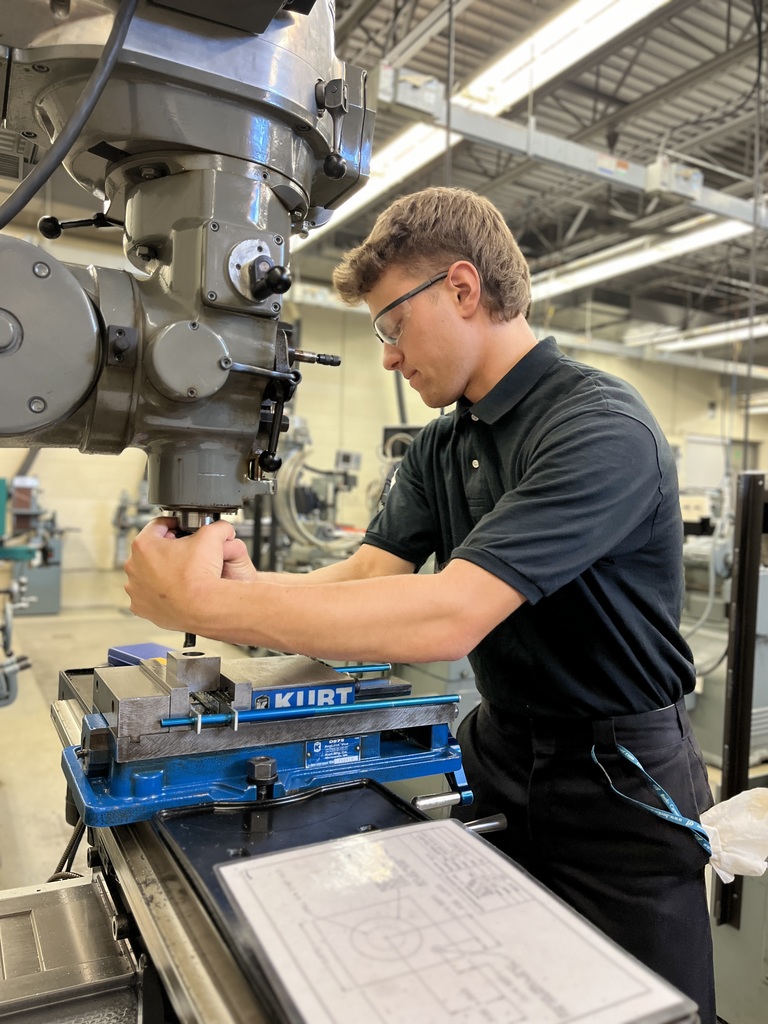 A person wearing safety glasses and a black polo shirt with the "CNC Machining" logo stands beside a large industrial milling machine in a brightly lit workshop. The machine features a blue vise labeled "Kurt" on its table, with additional equipment visible in the background.