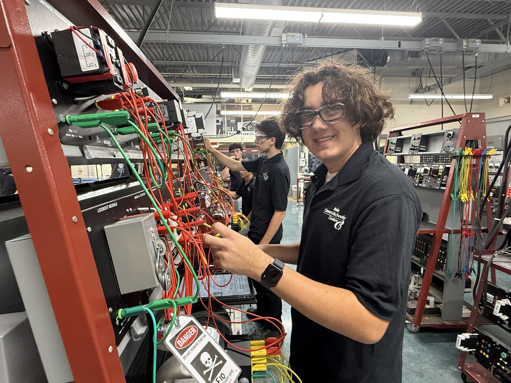 A group of students in a technical workshop are working on electrical wiring projects. In the foreground, a student with curly hair and glasses smiles at the camera while handling red and green wires connected to electrical components on a metal frame. Other students are visible in the background, also engaged in wiring tasks. The workshop features industrial lighting, equipment, and warning signs indicating electrical hazards.