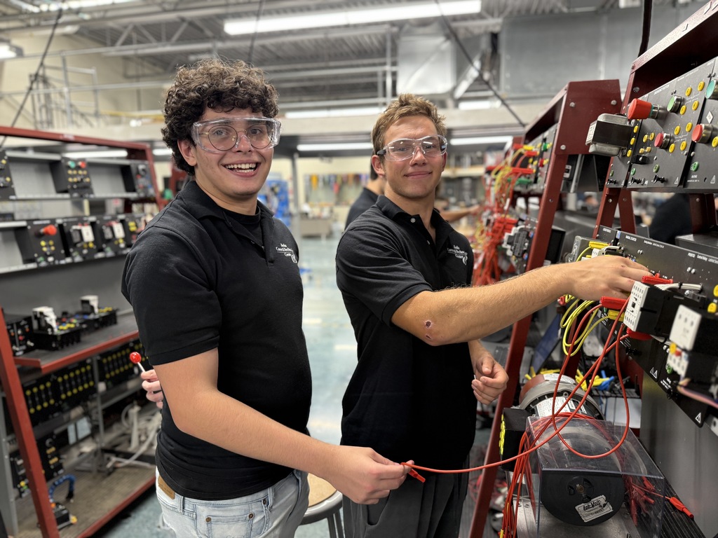 A person with curly hair and glasses is working on a large electrical panel filled with numerous red and green wires. The person is smiling at the camera while holding a wire. In the background, two other individuals are also working on similar panels. The setting appears to be an industrial or educational facility with various equipment and tools visible.