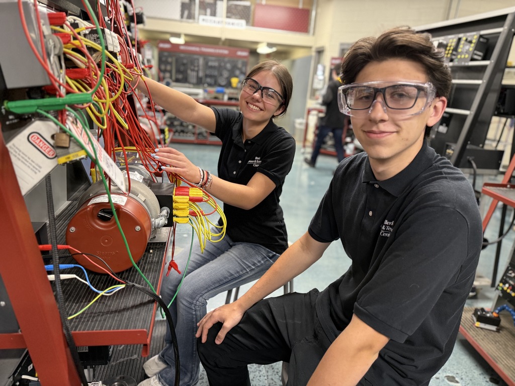 A person with curly hair and glasses is working on a large electrical panel filled with numerous red and green wires. The person is smiling at the camera while holding a wire. In the background, two other individuals are also working on similar panels. The setting appears to be an industrial or educational facility with various technical equipment visible.