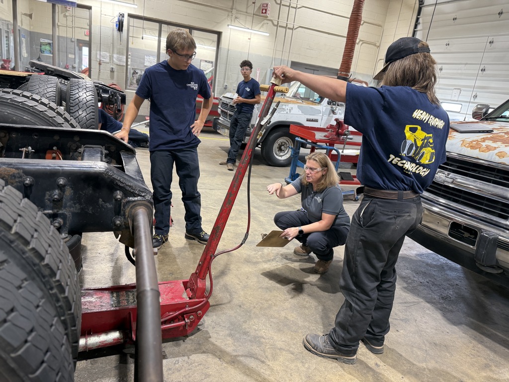 Four individuals are engaged in automotive repair in a workshop. One person operates a red hydraulic jack to lift part of a vehicle chassis, while another crouches nearby, possibly instructing or discussing the task. Tools, equipment, and vehicles are visible in the background, highlighting a collaborative, hands-on learning environment.