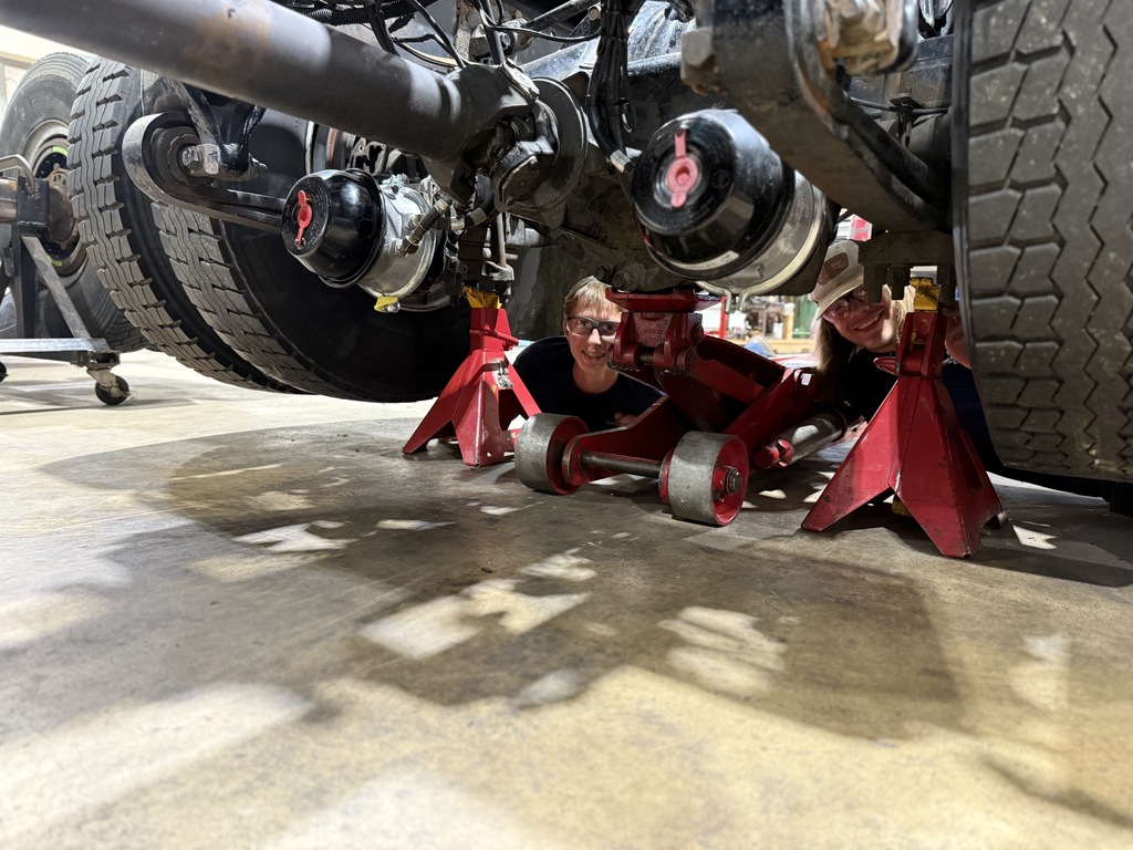 Two individuals are working beneath a truck supported by red jack stands and a hydraulic floor jack. The image captures the underside of the vehicle, emphasizing mechanical maintenance or inspection in progress.