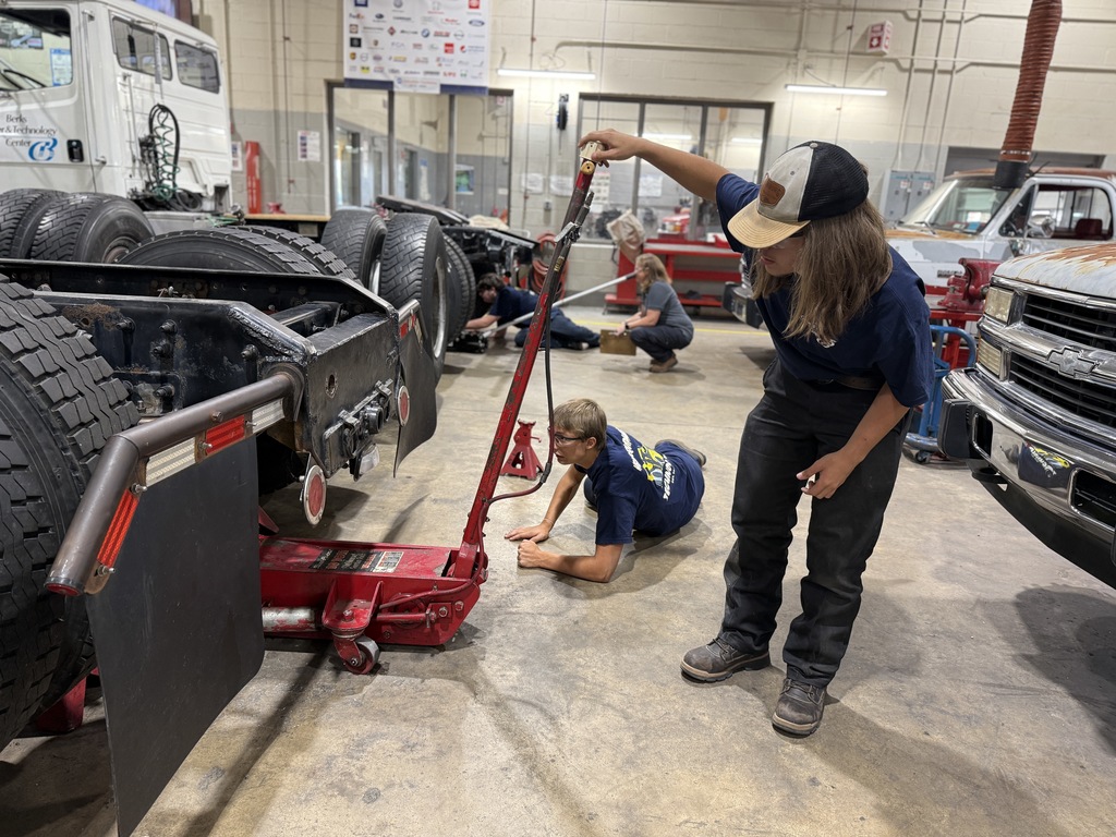 In an automotive workshop, a group of individuals are engaged in vehicle maintenance. One person operates a red hydraulic jack to lift the rear of a large vehicle, while another lies on the ground, possibly guiding the jack. Tools, equipment, and another vehicle are visible in the background, highlighting a collaborative repair environment.
