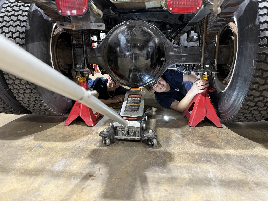 Two individuals lie on their backs beneath a vehicle elevated on red jack stands in a garage. They are working near the rear axle and differential, with a hydraulic floor jack positioned centrally. The concrete floor and surrounding tools suggest a workshop environment.