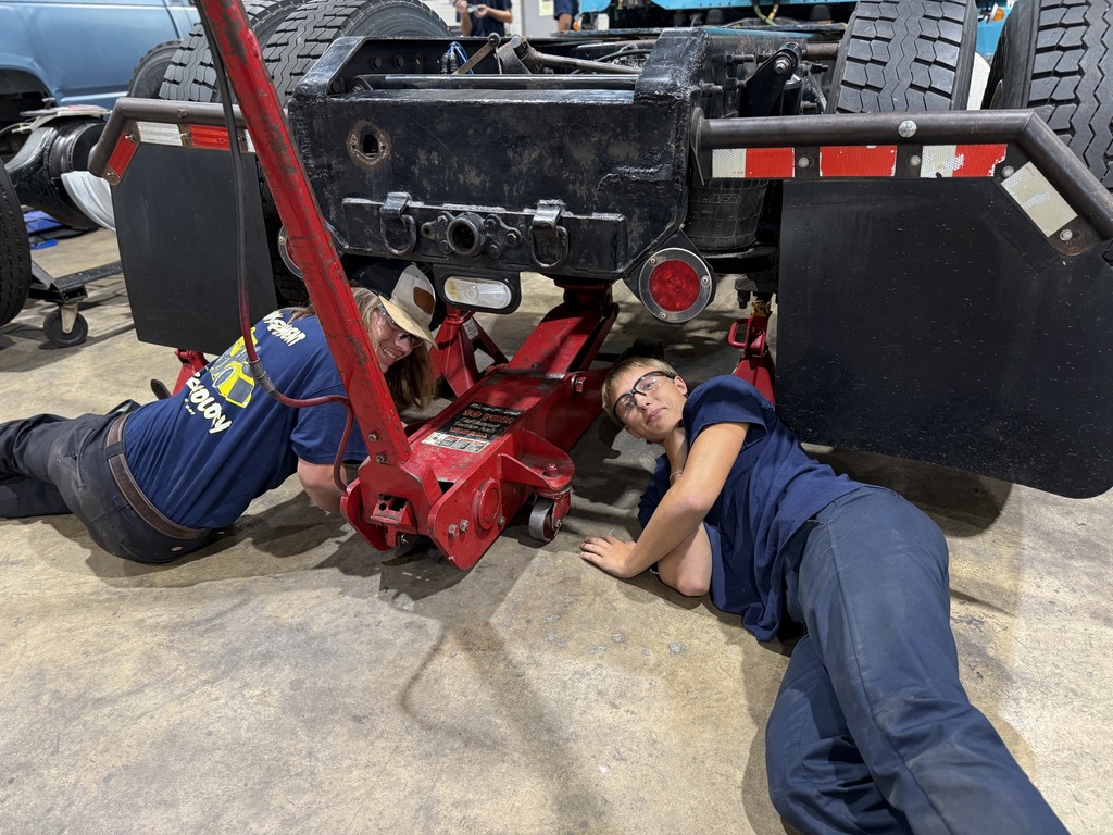 Two individuals are performing maintenance beneath a large vehicle, possibly a truck or trailer, using a red hydraulic floor jack to lift the rear. They are working on the concrete floor of an indoor garage, surrounded by tools, indicating a hands-on automotive repair setting.