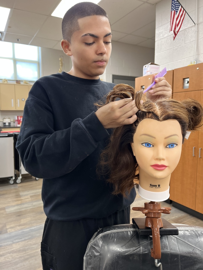 A Cosmetology student carefully parts and styles a mannequin head with medium brown hair. The mannequin is secured to a chair clamp, and the student uses a purple comb.