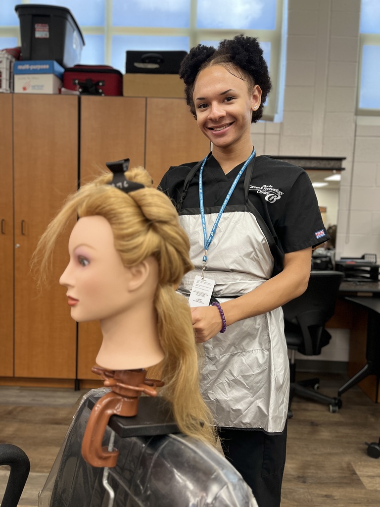 A Cosmetology student stands behind a mannequin head with sectioned blonde hair clipped up for styling. The student smiles while wearing a black uniform with a silver apron.