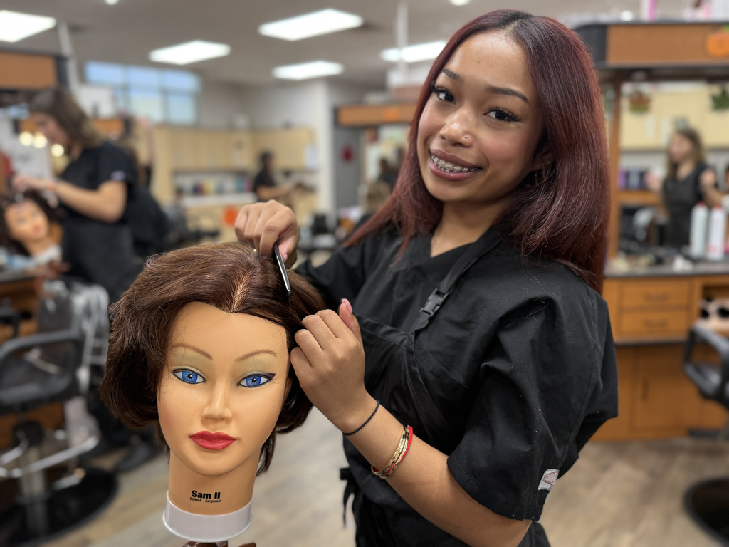 A Cosmetology student styles a mannequin head with straight brown hair. The student smiles at the camera while holding a comb.