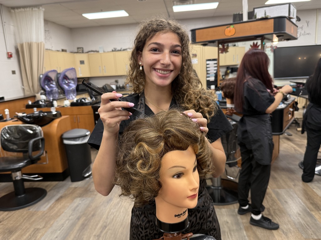 A Cosmetology student smiles while styling a mannequin head with short, curly light brown hair using a comb.