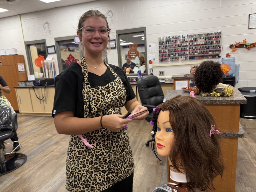 A Cosmetology student in a leopard-print apron styles a mannequin head with brown hair. The hair is sectioned with pink clips for curling.