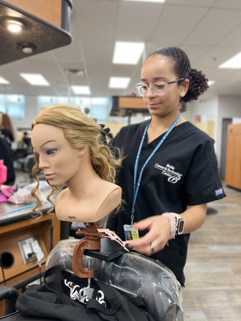 A Cosmetology student at Berks Career & Technology Center works on styling a mannequin head with long blonde hair secured to a chair clamp. The student is wearing a black uniform and glasses.