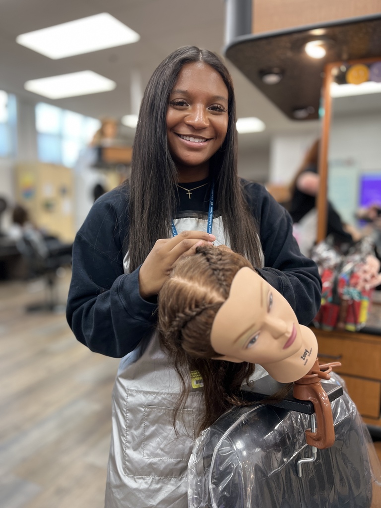 A smiling Cosmetology student braids the hair of a mannequin head in the classroom. The mannequin is tilted to the side, showing the braid in progress.