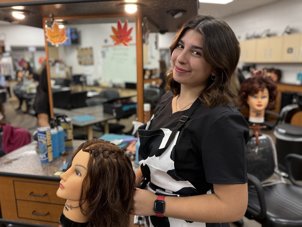 A Cosmetology student in a black and white apron braids the hair of a mannequin head. The student smiles while working in the classroom salon.