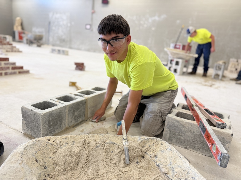 A student wearing safety glasses and a bright yellow shirt kneels beside a row of cinder blocks, carefully applying mortar with a trowel. A wheelbarrow filled with mortar and a red level rest nearby in the masonry lab.