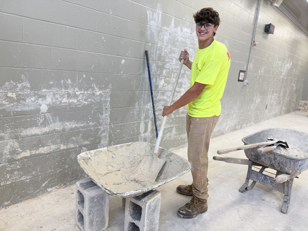 A student in a neon yellow shirt and safety glasses smiles while mixing mortar in a wheelbarrow with a long-handled tool inside the masonry shop.