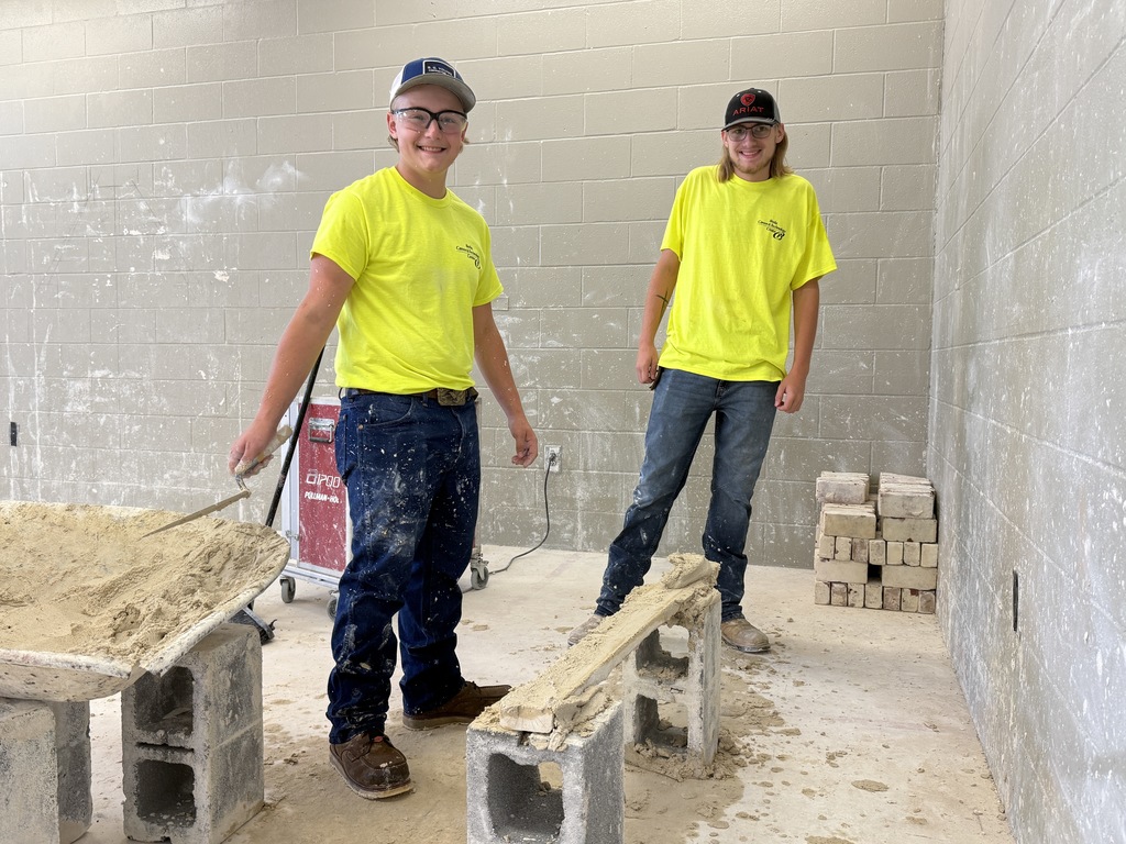 Two students wearing bright yellow shirts and safety glasses stand beside a wheelbarrow of mortar. One holds a trowel while smiling, and the other stands next to a short wall made of cinder blocks coated with fresh mortar.
