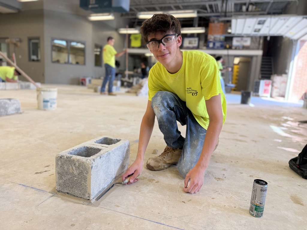 A student in a bright yellow shirt and safety glasses kneels on the shop floor, using a trowel to apply mortar to the base of a cinder block. A partially built wall and tools are visible in the background.