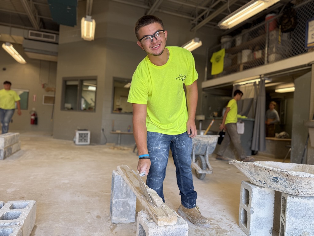A student in a neon yellow shirt and safety glasses applies mortar across the top of a row of cinder blocks using a trowel. The student smiles as they work in the masonry lab.