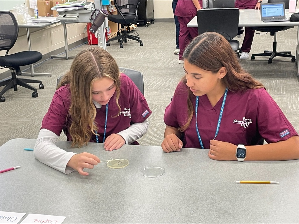 Two students in burgundy scrubs carefully observing a petri dish, one holding a swab to the dish