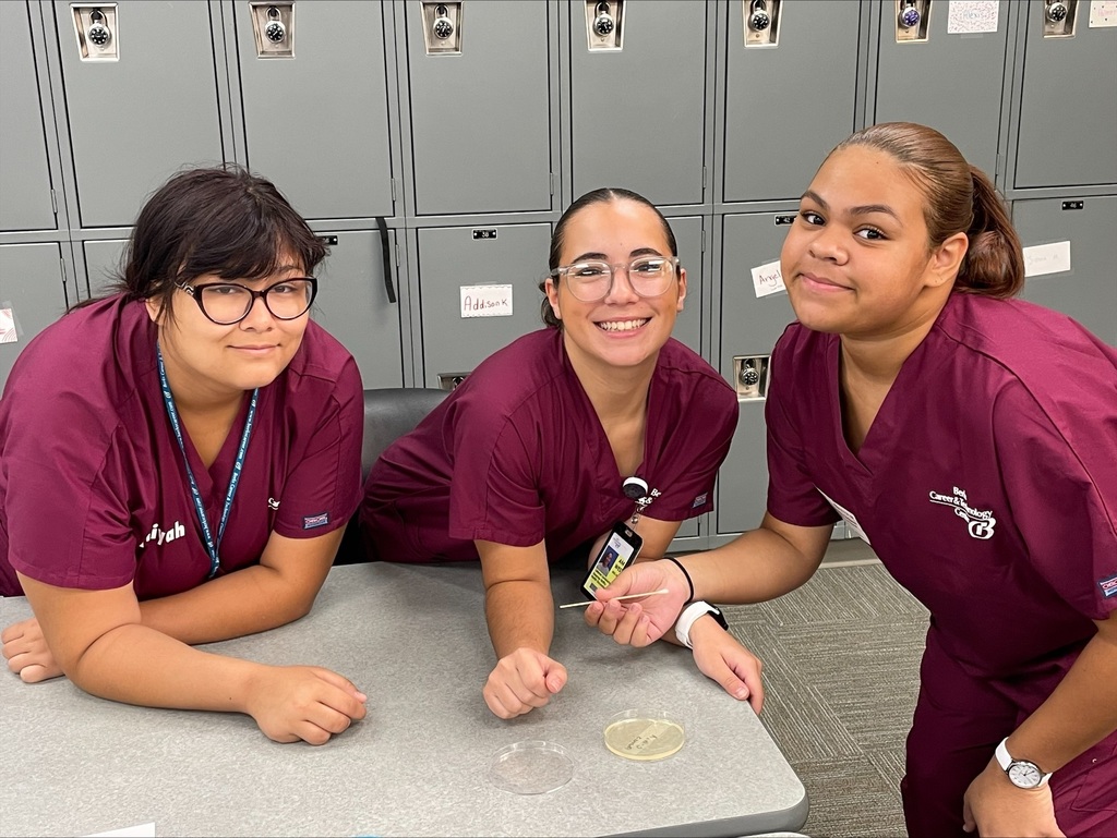 Three students in burgundy scrubs smiling at the camera with a petri dish on the table in front of them.