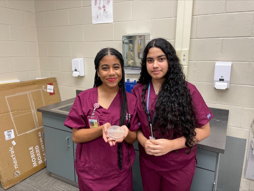 Two students in burgundy scrubs standing and holding a petri dish toward the camera with a smile.