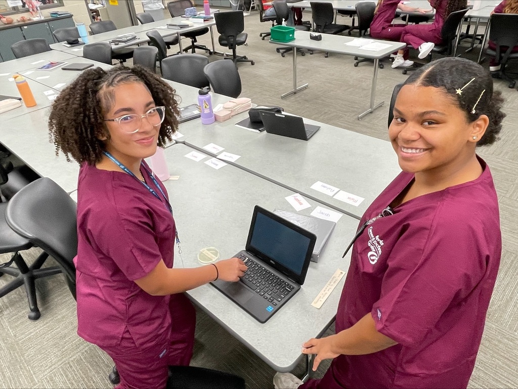 Two Health Occupations students in burgundy scrubs smiling at the camera while working on a laptop, with a petri dish nearby on the table.