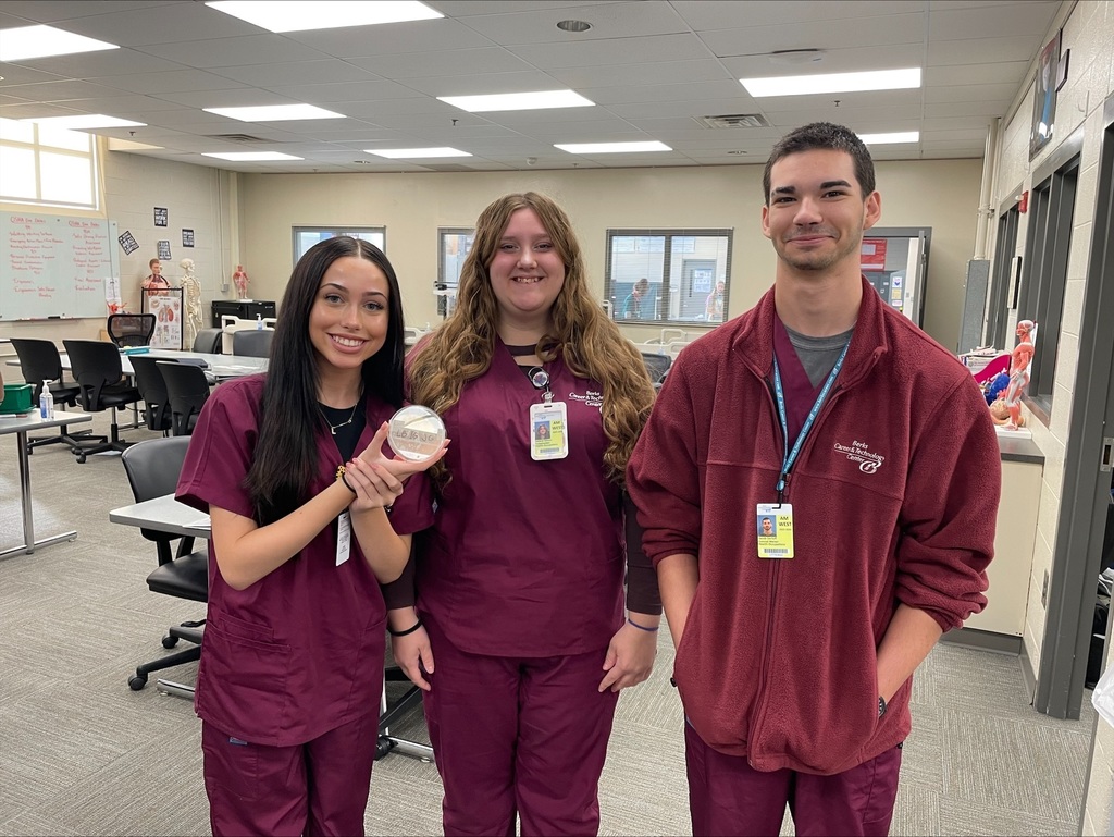Three students in burgundy scrubs standing in a classroom, smiling at the camera while one holds up a petri dish.