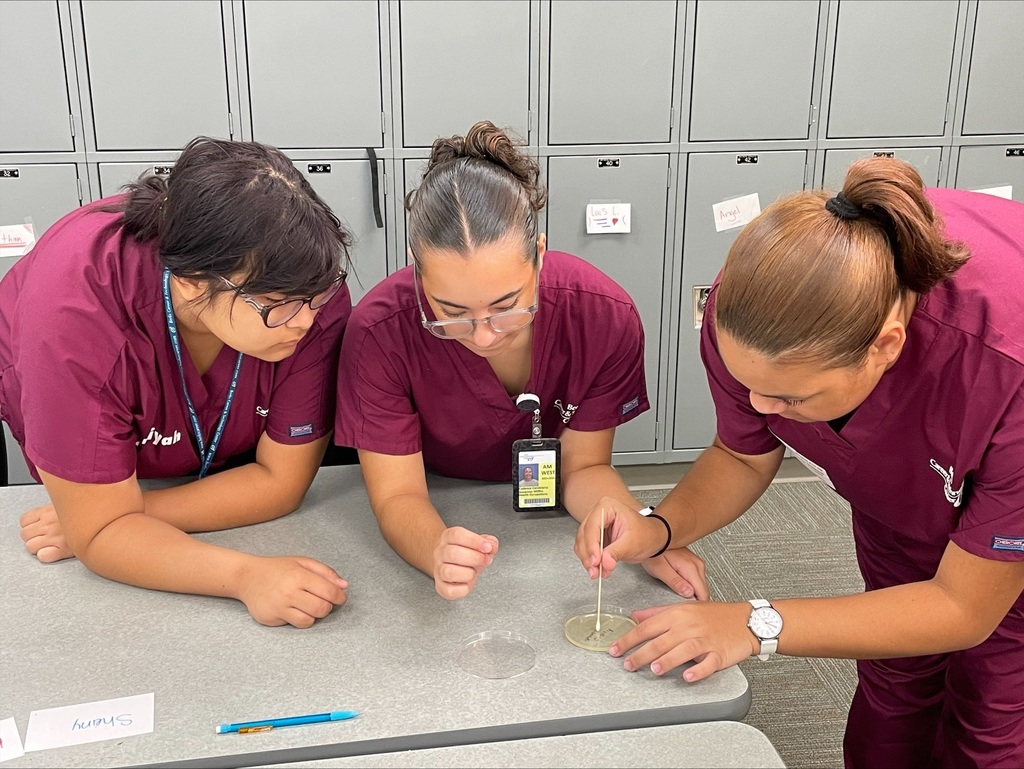 Three students in burgundy scrubs gathered around a table, concentrating on swabbing a petri dish.