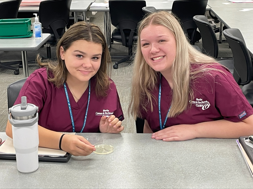Two students in burgundy scrubs smiling at the camera, one holding a petri dish on the table.