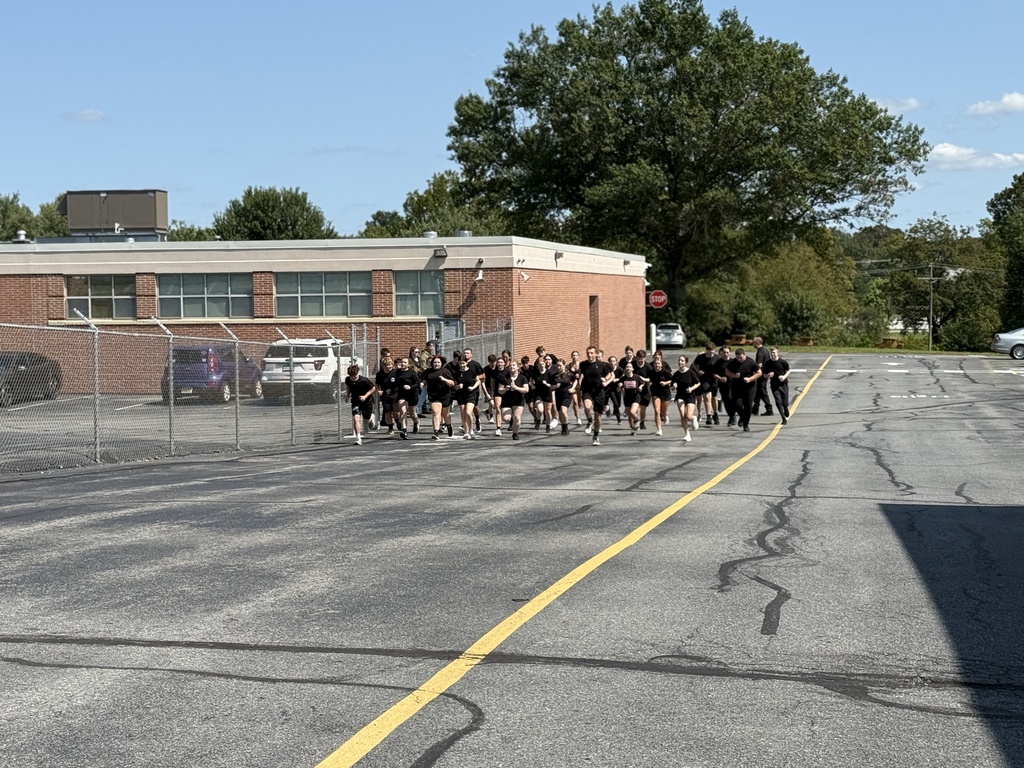 A large group of Protective Services students in black athletic uniforms running together on a paved lot outside the school building, starting a fitness exercise.