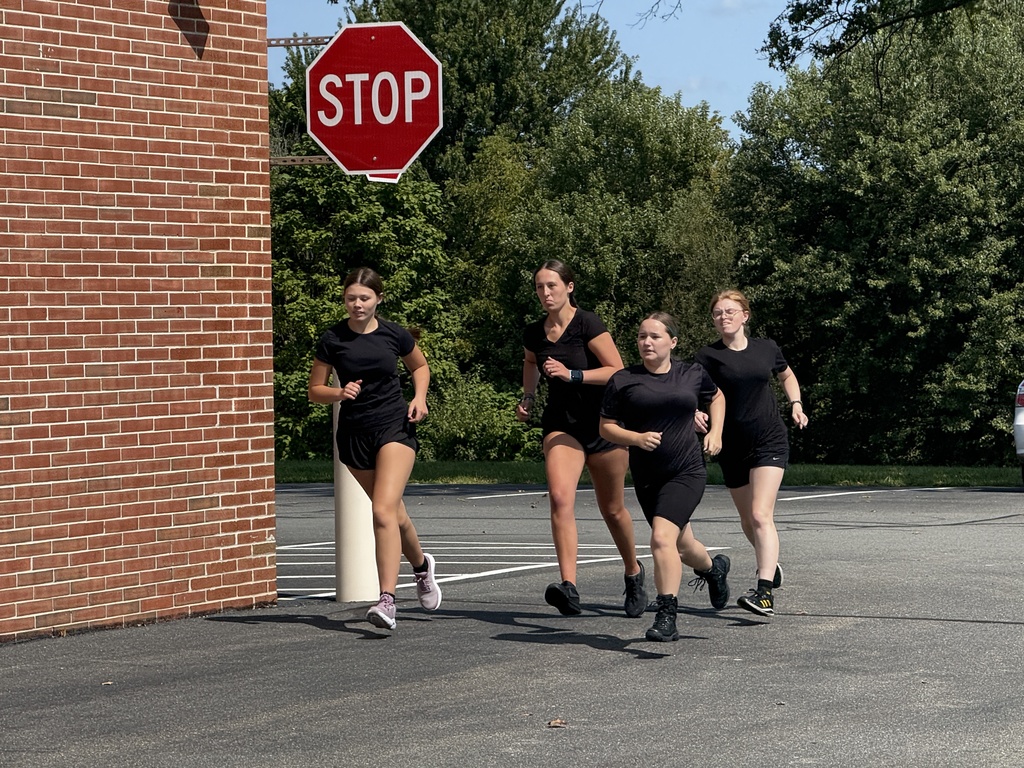 Four Protective Services students in black athletic uniforms running around the corner of a brick school building near a stop sign, focused on their pace.