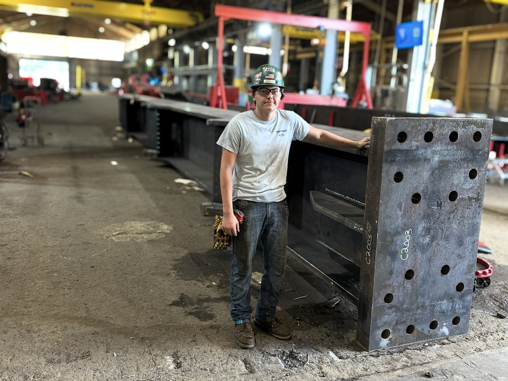 A person wearing a hard hat, safety glasses, light-colored shirt, jeans, and work boots stands in a large industrial workshop beside a massive steel beam with multiple holes drilled into its end. The workshop has high ceilings with yellow overhead cranes labeled "JGM" and an American flag hanging from one of them. The floor is scattered with tools and equipment, and the background includes machinery and workstations.