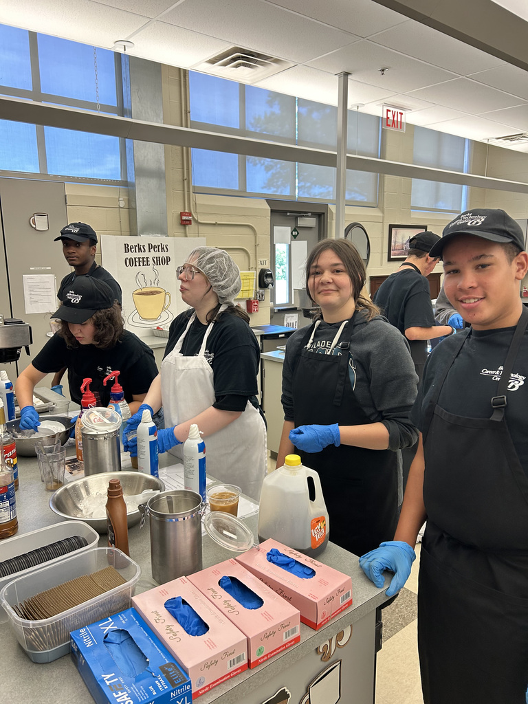 A group of individuals wearing black shirts, aprons, and gloves are working together in a kitchen setting. The counter is filled with kitchen items including mixing bowls, containers, milk, and syrup bottles. A sign in the background reads "Bert's Perks Coffee Shop," suggesting the activity is part of a coffee preparation or culinary training program.