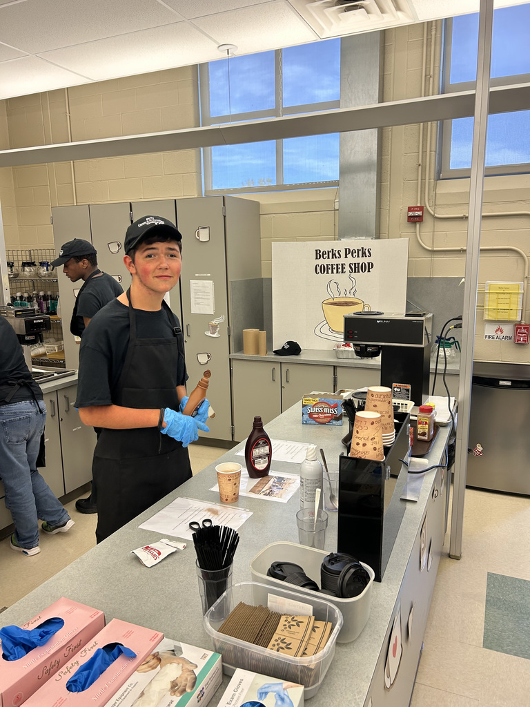 Two individuals in black uniforms and caps are working behind the counter of a coffee shop setup labeled "Berks Perks COFFEE SHOP." One person is holding a wooden rolling pin and wearing blue gloves. The counter displays coffee-making supplies including disposable cups, chocolate syrup, sweeteners, napkins, and straws. The background features cabinets and equipment, suggesting a clean and organized institutional or school-based environment.