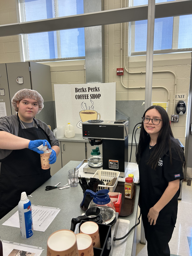 our individuals are in a classroom setting. In the foreground, two people wearing blue gloves stand side by side. The person on the left, in a gray sweatshirt, is smiling and holding a cup with brown liquid. The person on the right wears a yellow fleece jacket with an ID badge and has hands resting on the table. The table holds boxes of gloves, containers, and papers. In the background, two other individuals are visible—one facing away and another near computers.