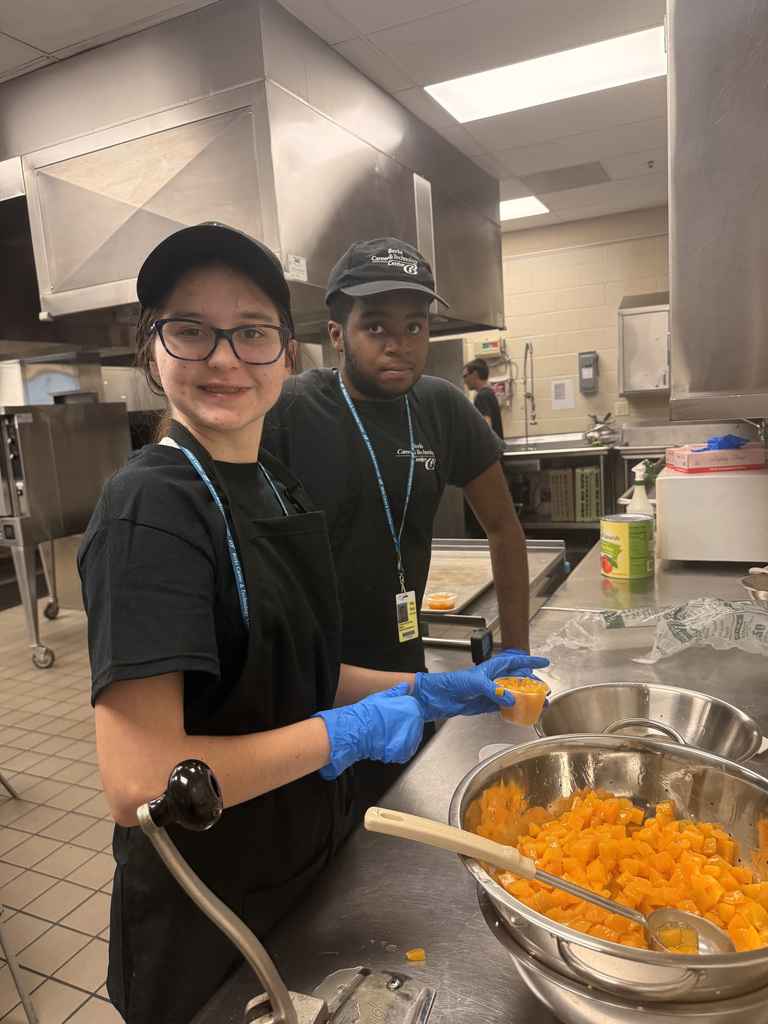 Two individuals in black uniforms, hats, aprons, and blue gloves are working in a commercial kitchen. One person holds a small container of diced orange fruit, while the other stands beside a large metal bowl filled with the same fruit. The kitchen features stainless steel appliances and countertops, indicating a professional food preparation environment.