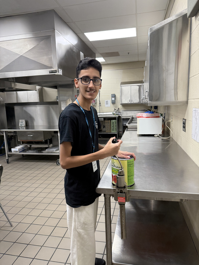 A person is standing in a commercial kitchen using a mounted can opener to open a large can labeled "S&W Organic Black Beans." The kitchen includes industrial equipment such as ovens and stainless steel cabinets, and appears clean and organized, suggesting a professional food preparation environment.