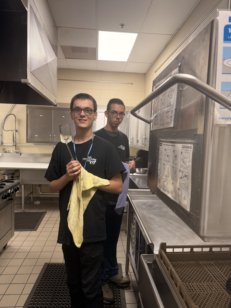 Two individuals are working in a commercial kitchen. The person in the foreground holds a spatula and a yellow cloth, while the person in the background holds a blue cloth. Both wear black shirts with logos and appear to be engaged in cleaning tasks. The kitchen includes stainless steel appliances, sinks, countertops, and tiled floors and walls, indicating a professional food service environment.