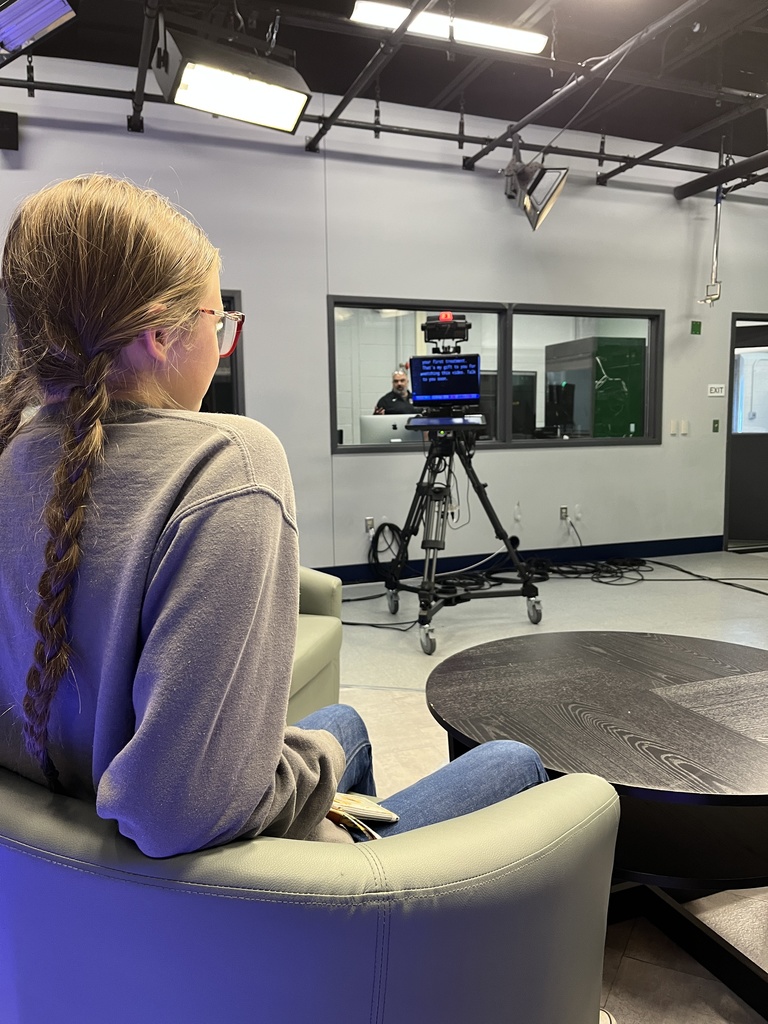 A student with a long braid sits in a studio chair, facing a teleprompter mounted on a camera. The teleprompter shows white text on a black background, with studio lights and monitors visible in the room.
