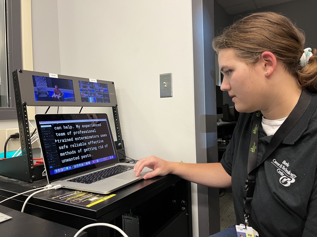 A student in a black Berks Career & Technology Center polo shirt uses a laptop to edit teleprompter text. The screen shows large white words on a black background as the student focuses on the setup.