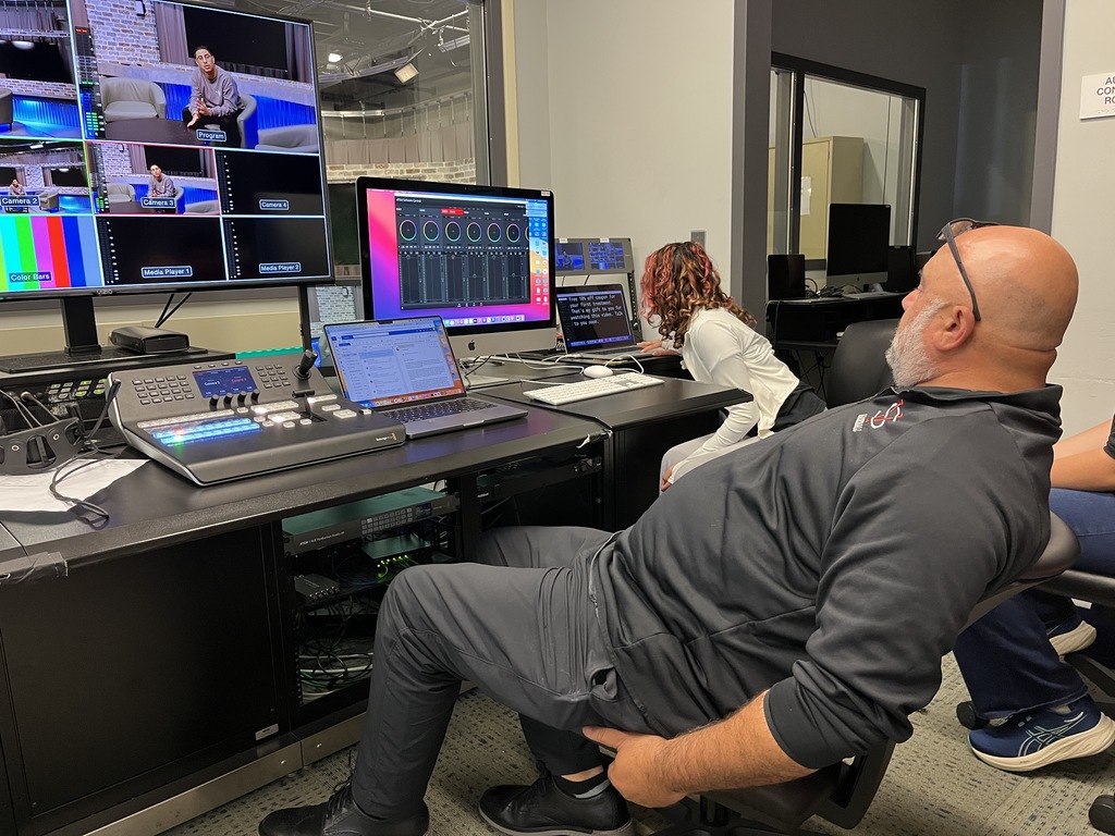 An instructor and students work in a production control room. Multiple monitors display live video feeds and sound levels while a student adjusts a teleprompter script on a computer.