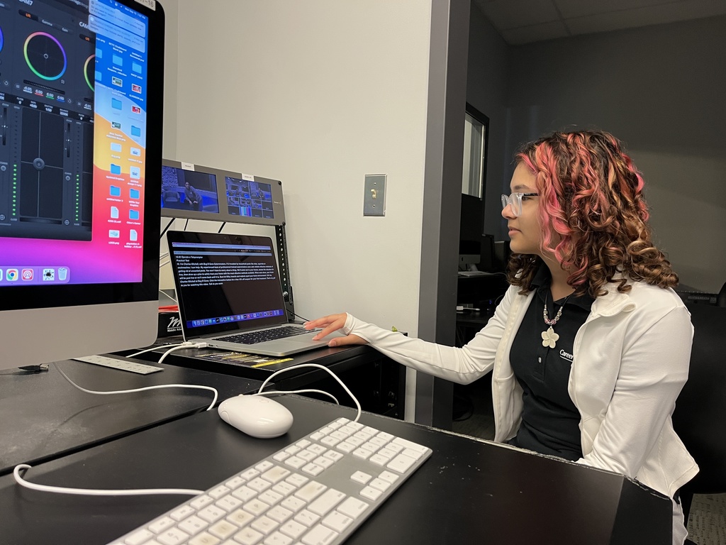 A student wearing glasses and a white jacket operates a laptop displaying a teleprompter script. Studio control room monitors and equipment are visible around the student.