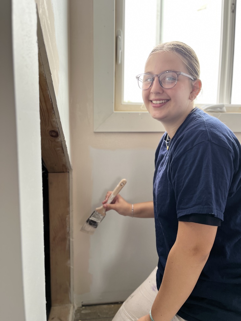 An individual painting an indoor wall with a brush. They are wearing glasses, a dark blue shirt, and white pants. The wall is partially painted, and natural light enters through a nearby window.