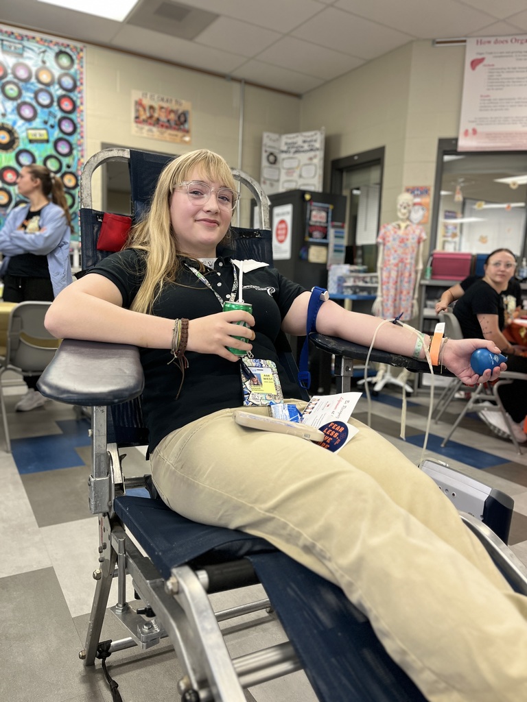A BCTC student with blond hair and glasses donates blood while seated in a chair. She holds a green drink cup and a blue stress ball, smiling slightly at the camera.