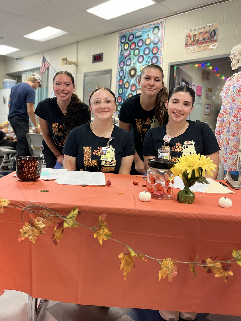 Four Health Occupations students from Tulpehocken smile at a decorated sign-in table for the blood drive. The table is covered with an orange cloth, small pumpkins, a sunflower centerpiece, and autumn garland.