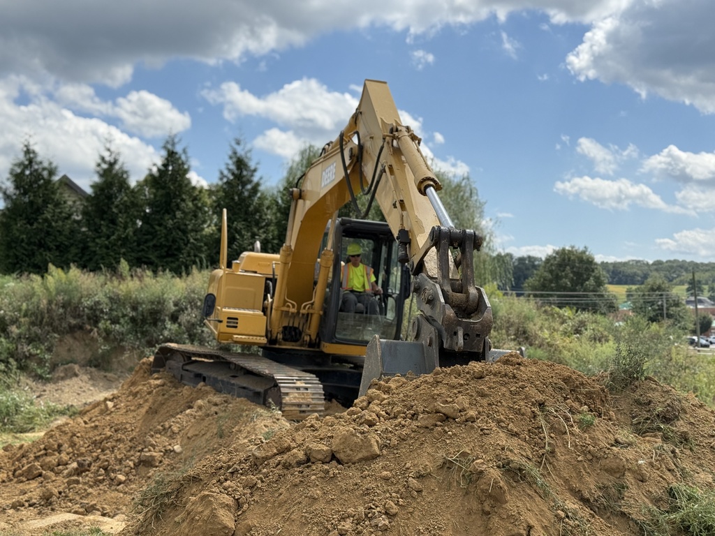 A close-up view of a John Deere excavator as a student in a safety vest and helmet operates it while digging into a dirt mound.